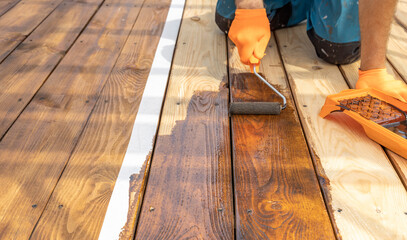 A person wearing orange gloves is using a roller to apply stain to wooden deck boards. The deck...