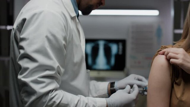 Side shot of bearded immunologist in lab coat and gloves doing injection or vaccination, inserting needle into arm of female patient in modern clinic