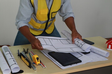 An engineer meticulously draws house plans on a workbench, surrounded by tools and blueprints,...