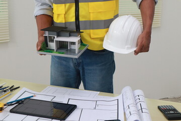 An engineer meticulously draws house plans on a workbench, surrounded by tools and blueprints,...