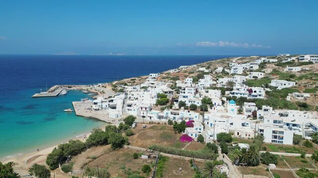 An aerial perspective reveals a stunning view of Donousa in the Cyclades, Greece. Whitewashed buildings cascade down a hillside, overlooking a turquoise bay with a sandy beach and a small harbor.