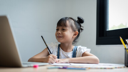 A young girl is sitting at a desk with a laptop and a pencil. She is smiling and she is enjoying herself. The scene suggests that she is engaged in a creative activity, such as drawing or writing