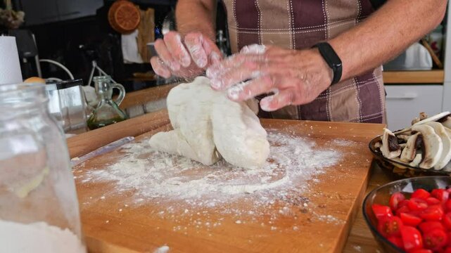 Man kneads pizza dough on a wooden surface. The background features a home kitchen, suggesting he's making dinner for his family. Natural lighting in a real kitchen environment.