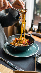 A person is twirling pasta with a fork in a black bowl, garnished with vegetables. The setting is a cozy restaurant with natural light, perfect for culinary and dining content.