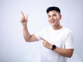 A young Asian man, wearing a white shirt and black watch, smiles widely while pointing both index fingers to his left. The background is plain white, highlighting the subject's joyful