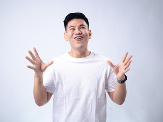 Fototapeta premium A young Asian man, wearing a white shirt and black watch, has a surprised and exaggerated expression with his mouth open and hands spread wide. The plain white background highlights