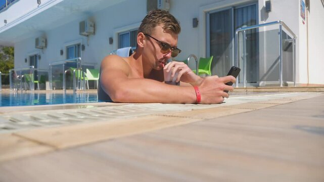 Man using phone at pool and refreshing cold cocktail. Guy drink beverage browsing internet page or communicating with somebody. Young man enjoying recreation on resort during summer vacation. Low view