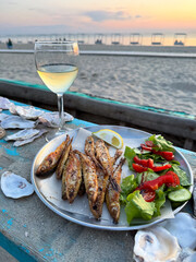 Plate of grilled fish with fresh salad and a glass of white wine on a beachside table at sunset, perfect for a relaxing evening.