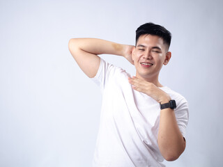 A young Asian man, wearing a white shirt, makes a disgusted face while smelling his armpit. The plain white background emphasizes the subject's humorous and exaggerated reaction