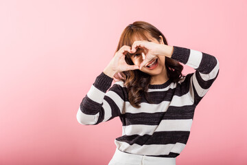 An Asian lady smiles confidently while forming a heart symbol with her fingers and hands on a pink background. A beautiful portrait of a young woman expressing love and happiness.