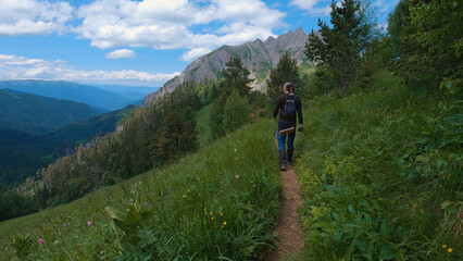 Fototapeta premium A man is walking along a narrow path in the mountains with majestic mountains looming in the background. The sky is clear, with only a few clouds adding to the tranquility.