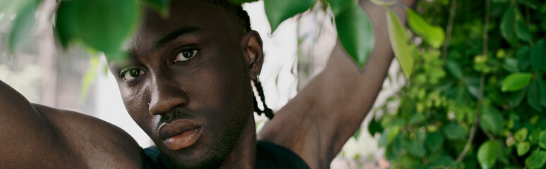 Handsome African American man with dreadlocks poses in front of a lush tree in a green garden.