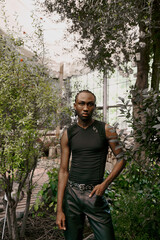 An African American man with dapper style stands gracefully in front of lush green trees.