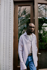 Handsome African American man with dapper style stands confidently in front of a door in a vivid green garden.