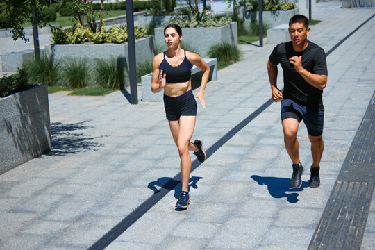 Man and woman in sports attire running side by side. Bright sunlight and their energetic pace convey sense of vitality and health. Concept of sport, healthy lifestyle, pair training, leisure activity.