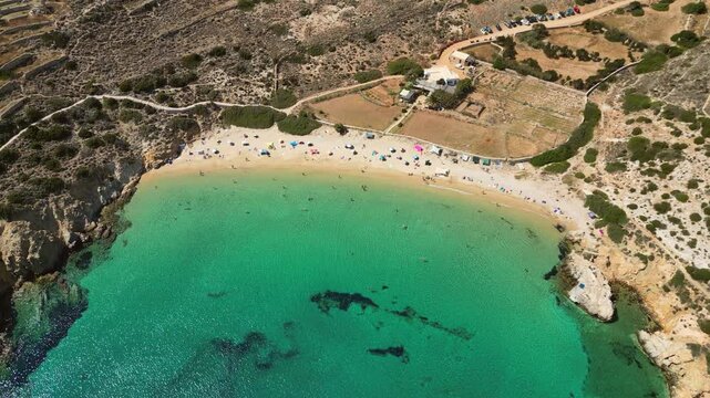 A picturesque view from above of Kedros Beach on the Greek island of Donousa, featuring clear turquoise waters, golden sand, and a small number of people enjoying the sun and sea.