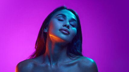 Femininity and sensuality. Attractive elegant young women with bare shoulders, long dark hair against purple background in neon light. Concept of beauty, skin and body care, cosmetology and cosmetics