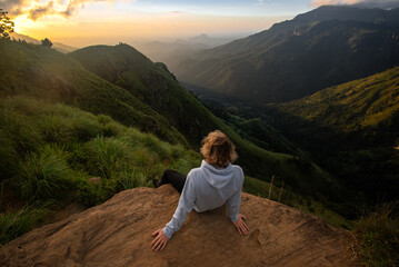 Naklejka premium A woman sits on a rock amidst the rugged terrain of the Sri Lankan mountains, taking in the panoramic view.