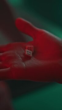 Vertical closeup of hand of unrecognizable person opening palm and holding two ecstasy pills with emoticon in nightclub with red and green neon light