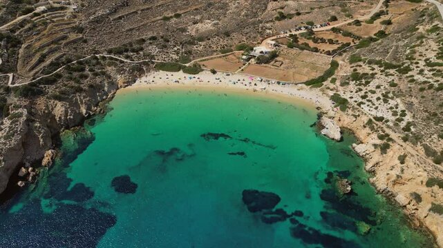 An aerial view of Kedros Beach on Donousa Island in the Cyclades, Greece. The crystal-clear turquoise waters lap against the golden sands, dotted with sunbathers and umbrellas.