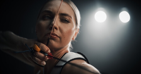 Close-Up Portrait Of Professional Female Archer in Action. Portrait of Beautiful Athlete Holding a Bow and Pulling Back the Arrow in Order to Hit the Target in a Dark Competition Room