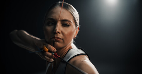 Portrait of a Beautiful Female Archer Holding a Modern Compound Bow, Aiming at a Target. Young Athlete Drawing the Arrow in a Stadium Competition or in a Dark Studio for Advertising Online