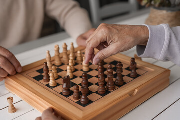 Close up on chess board and hands of senior couple of people playing at home with chess on wooden...