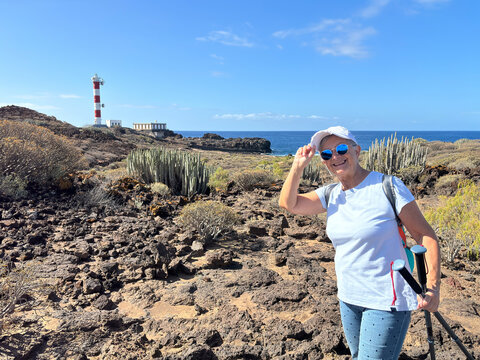 Outdoor activity in nature for a carefree sporty senior woman walking along a rocky beach in direction to a lighthouse enjoying healthy lifestyle and freedom in good sunny day with blue sky