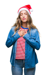 Beautiful young blonde woman wearing christmas hat over isolated background smiling with hands on chest with closed eyes and grateful gesture on face. Health concept.