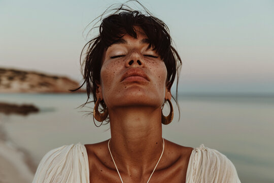 A person stands by the calm ocean shore at sunset, wearing gold hoop earrings and a white top, creating a perfect blend of elegance and natural beauty in a serene environment.