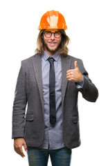 Young handsome architec man with long hair wearing safety helmet over isolated background doing happy thumbs up gesture with hand. Approving expression looking at the camera with showing success.