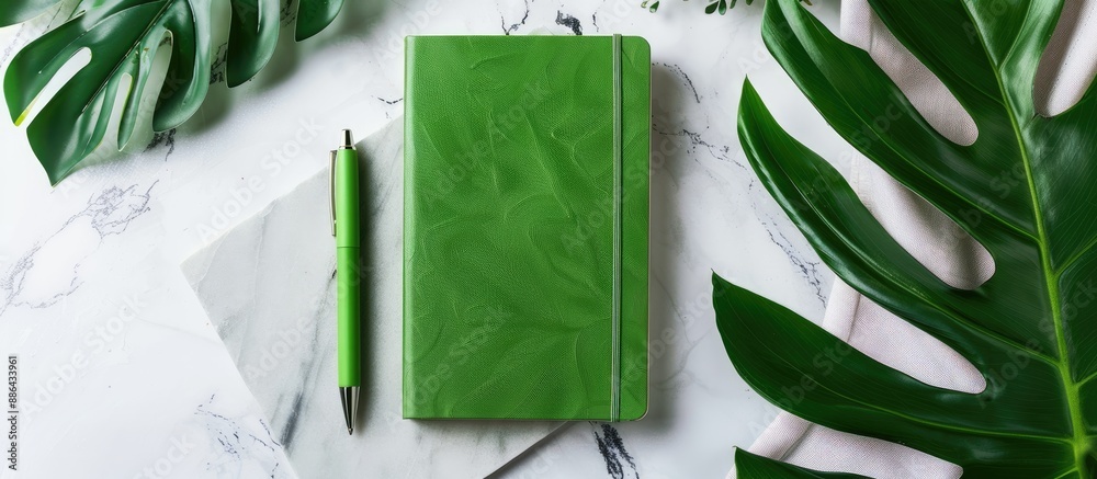 Poster a green notebook and pen on a white marble surface surrounded by a monstera leaf symbolizing busines
