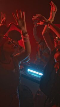 Vertical lowkey shot of group of young energetic ravers in retro y2k outfits waving hands and dancing to electronic music, hanging out in club with red neon light and flashes at night