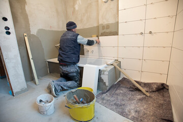 In einem Neubau klebt ein Bauarbeiter und Fliesenleger neue Fliesen an die Wand von einem Badezimmer