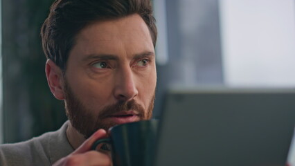 Closeup guy drinking coffee reading tablet at morning apartment. Man holding tab