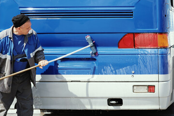 Uniform-clad individual thoroughly washes the back side of a bus, focusing on the taillights area