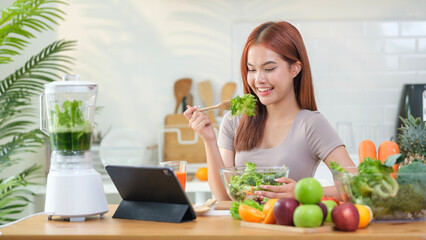 Young woman eating healthy vegetable salad and using digital tablet on the kitchen table with green fresh ingredients.