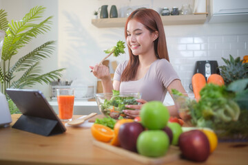 Young woman eating healthy vegetable salad and using digital tablet on the kitchen table with green fresh ingredients.