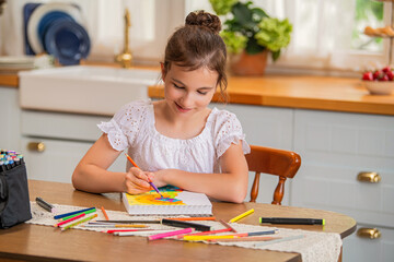 a girl draws in an album with colored pencils while sitting at a table in the interior of a country house kitchen.