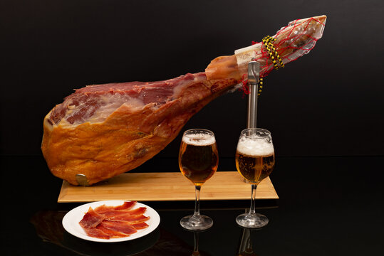 Smoked jamon meat on a metal stand holder, a plate with cold cuts and glasses with beer. Spanish traditional food, and an appetizer for beer on a black background in a restaurant. meat product