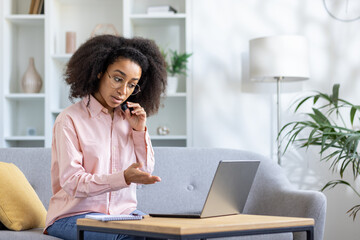 Obraz premium Young woman working from home talking on phone while using laptop. She appears to be explaining something during call. Modern home office setup on cozy couch in bright living room.