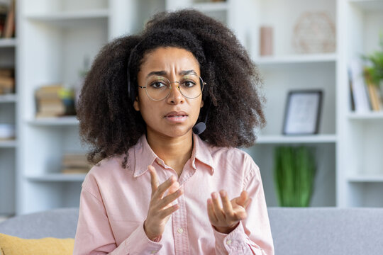 Woman wearing headset sitting at home office, engaging in conversation with frustrated expression on face, gesturing hands. Bookshelves and plants in background, creating professional environment. - Powered by Adobe