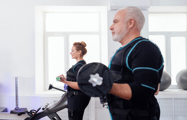 Older smiling couple in ems suits doing electrical muscle stimulation workout with dumbbells in gym or fitness club