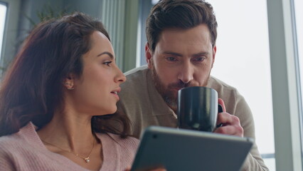 Morning pair looking tablet screen talking at grey panoramic apartment closeup