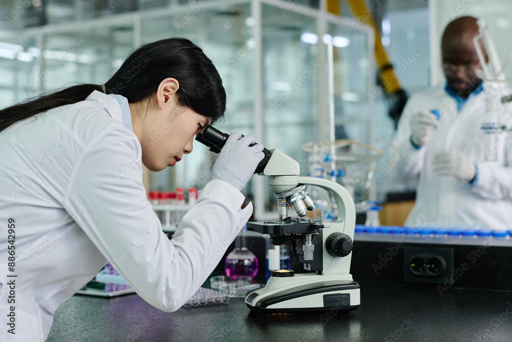Poster side view of young chinese female biochemist in whitecoat looking in microscope while studying new v