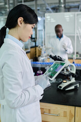 Fototapeta premium Young Chinese female scientist in gloves and lab coat pointing at screen of tablet while studying new virus or strain in laboratory