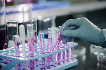 Gloved hand of modern chemist or clinician putting sample of purple liquid substance in stand with flasks during laboratory work