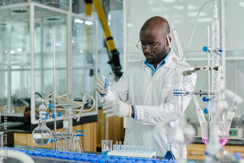 Poster young african american scientist in lab coat standing by workplace with glassware and carrying out e