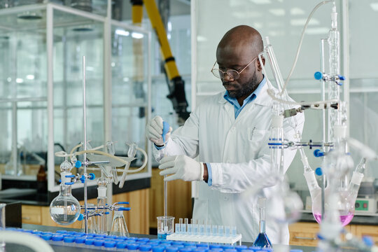 Young African American scientist in lab coat standing by workplace with glassware and carrying out experiment with liquid substances