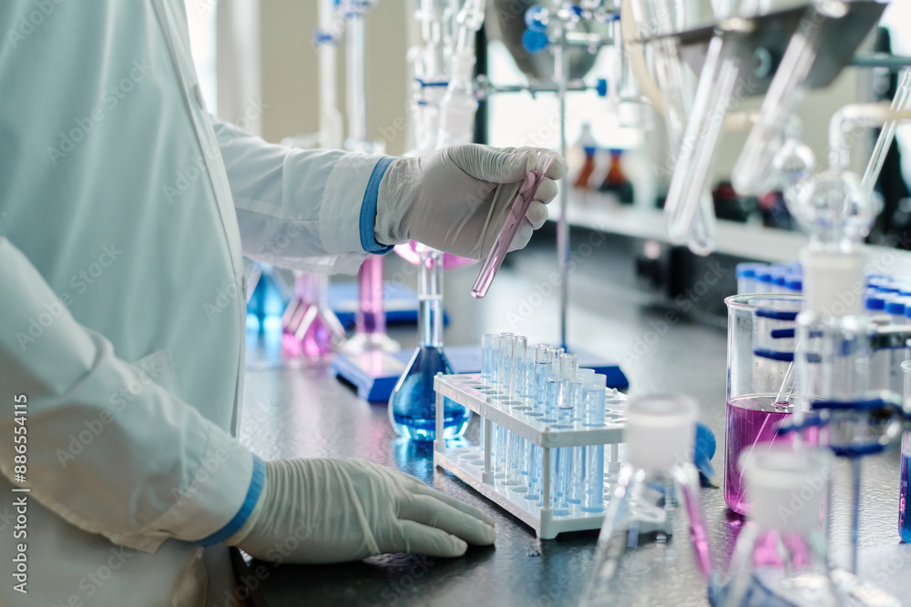 Poster Gloved hand of modern researcher or chemist in lab coat holding flask with liquid substance over set of samples during experiment